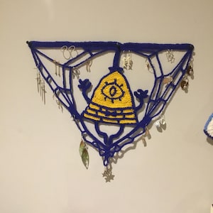 Puede incluir: Un tapiz de pared de crochet azul y amarillo con forma triangular y un ojo de dibujos animados en el centro. El tapiz tiene varios pendientes de plata colgando.