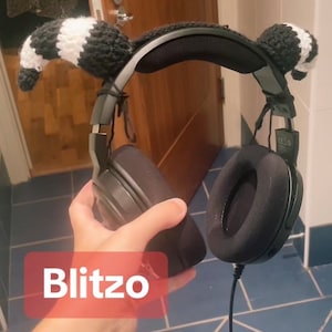 Könnte beinhalten: Schwarz-weiße gehäkelte Waschbär-Ohren-Abdeckungen, die an einem schwarzen Gaming-Headset mit einem schwarz-grauen Kopfbügel befestigt sind. Das Headset hat schwarze Ohrmuscheln mit einer weichen, schwarzen Polsterung. Der Text "Blitzo" ist im Bild sichtbar.