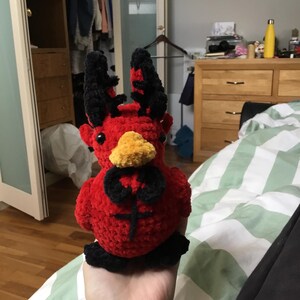 May include: A red and black crocheted plush toy with horns and a beak. The toy is being held in a person's hand.