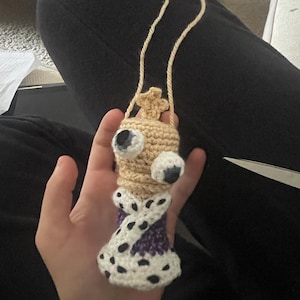 May include: A handmade crochet doll with a unique design. The doll features a beige head with large, googly eyes, a crown, and a twisted body in purple and white with black spots. A beige cord is attached for hanging.