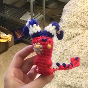 May include: A handmade crochet creature in red, white, and blue. The creature has a red body, white and blue striped ears, and a tail with blue stripes. A small white flower with a yellow center is on the front.