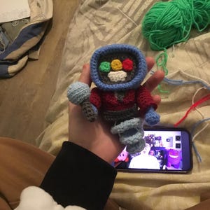 Pode incluir: Uma figura de crochê feita à mão que se assemelha a uma televisão retrô. A figura tem uma moldura azul, um corpo vermelho e uma base cinza. A tela apresenta círculos coloridos e um rosto sorridente. Um microfone azul claro está anexado.