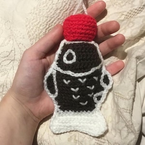 May include: A handmade, crocheted fish ornament. The fish is black with white detailing and a white tail. A red crocheted ball is attached to the top. The ornament is held by a hand.