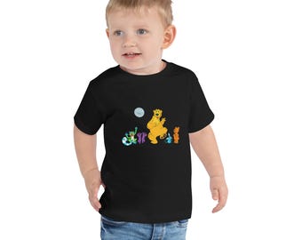 May include: Black toddler t-shirt with a colourful graphic of cartoon characters, including a yellow bear, a blue and green creature, and two orange figures. The design also includes a moon. The shirt is worn by a young child.