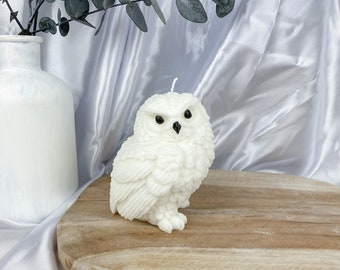 May include: A white owl-shaped candle with detailed feather textures. The candle sits on a wooden cutting board against a white satin backdrop. A white vase with eucalyptus branches is in the background.