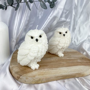 May include: Two white owl-shaped candles with black eyes and beaks sit on a wooden board. The candles have detailed feather textures and are lit with white wicks. The background is a white satin fabric.