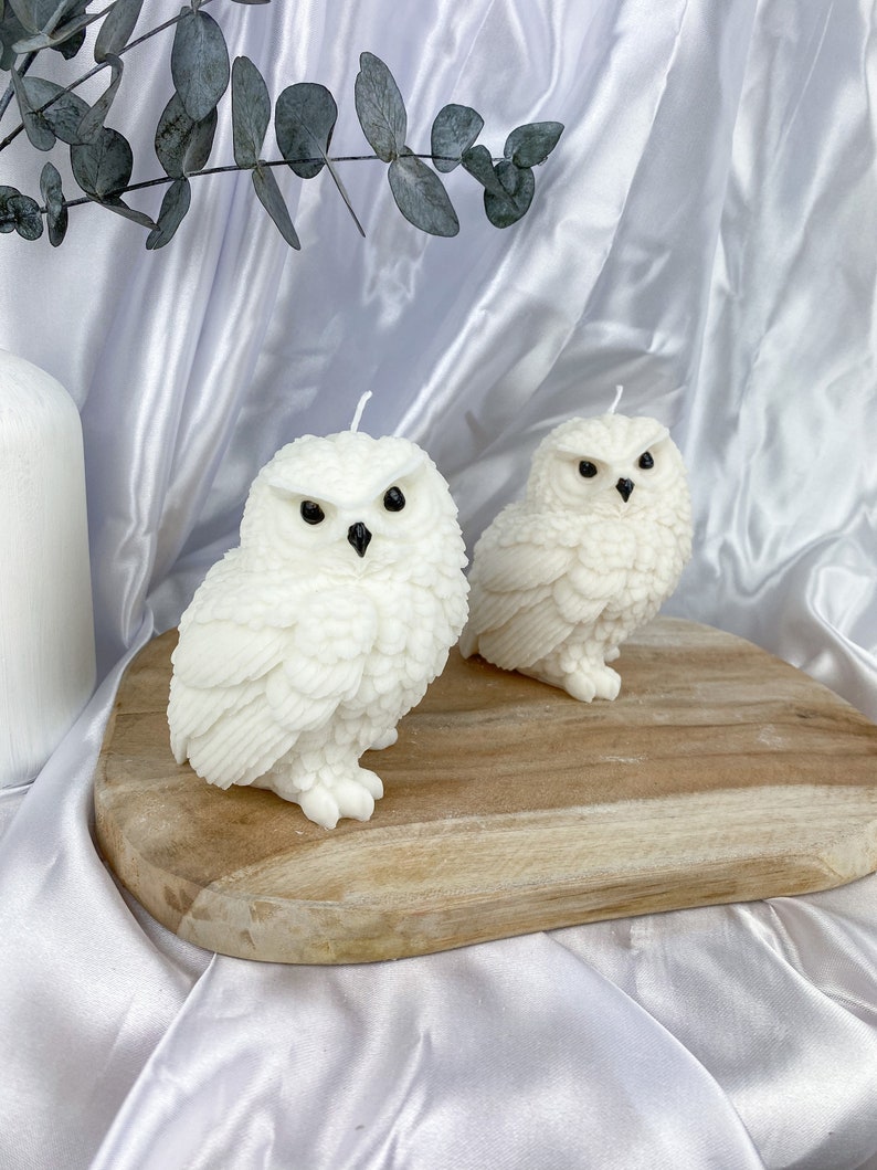 May include: Two white owl-shaped candles with black eyes and beaks sit on a wooden board. The candles have detailed feather textures and are lit with white wicks. The background is a white satin fabric.