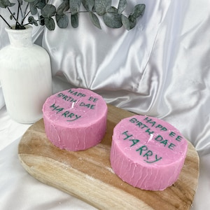 May include: Two pink birthday candles with the text "Happy Birthday" and "Harry" written on them. The candles are on a wooden board.