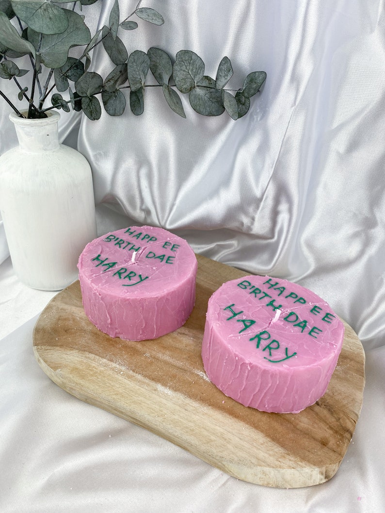 May include: Two pink birthday candles with the text "Happy Birthday" and "Harry" written on them. The candles are on a wooden board.