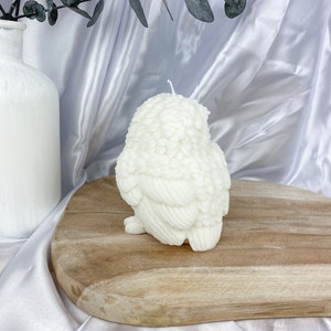 May include: A white owl-shaped candle with detailed feather textures. The candle sits on a wooden board against a white satin backdrop. A white vase with eucalyptus branches is visible in the background.