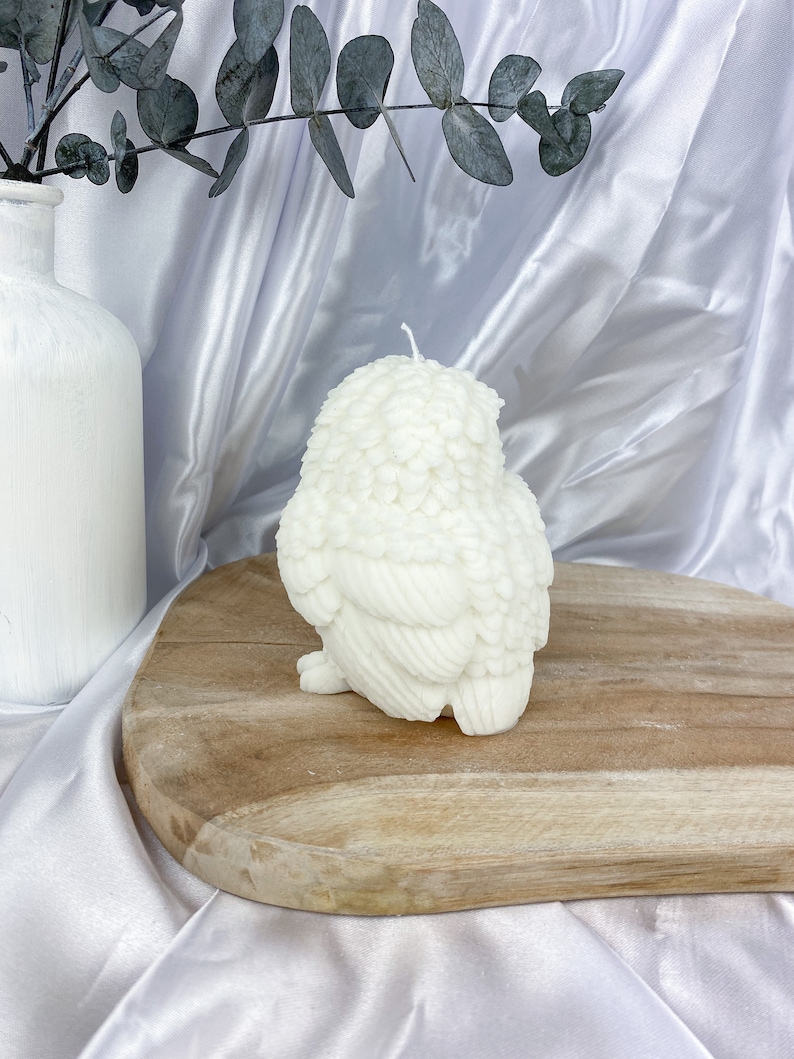 May include: A white owl-shaped candle with detailed feather textures. The candle sits on a wooden board against a white satin backdrop. A white vase with eucalyptus branches is visible in the background.