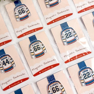 May include: Multiple magnetic bookmarks featuring white hockey jerseys with blue and orange accents. Each jersey displays a player name and number, including Logan 22, Di Laurentis 66, and Graham 44. The bookmarks are attached to individual display cards.