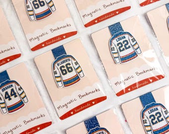May include: Multiple magnetic bookmarks featuring white hockey jerseys with blue and orange accents. Each jersey displays a player name and number, including Logan 22, Di Laurentis 66, and Graham 44. The bookmarks are attached to individual display cards.