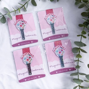 May include: Four magnetic bookmarks featuring a black lightstick design with a circular top containing colorful cartoon characters and the BTS logo. Each bookmark is attached to a pink card labeled Arirang Magnetic Bookmarks handmade.