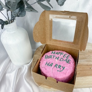 May include: A pink cake with green icing that says "Happy Birthday Harry" in a cardboard box with a clear window.