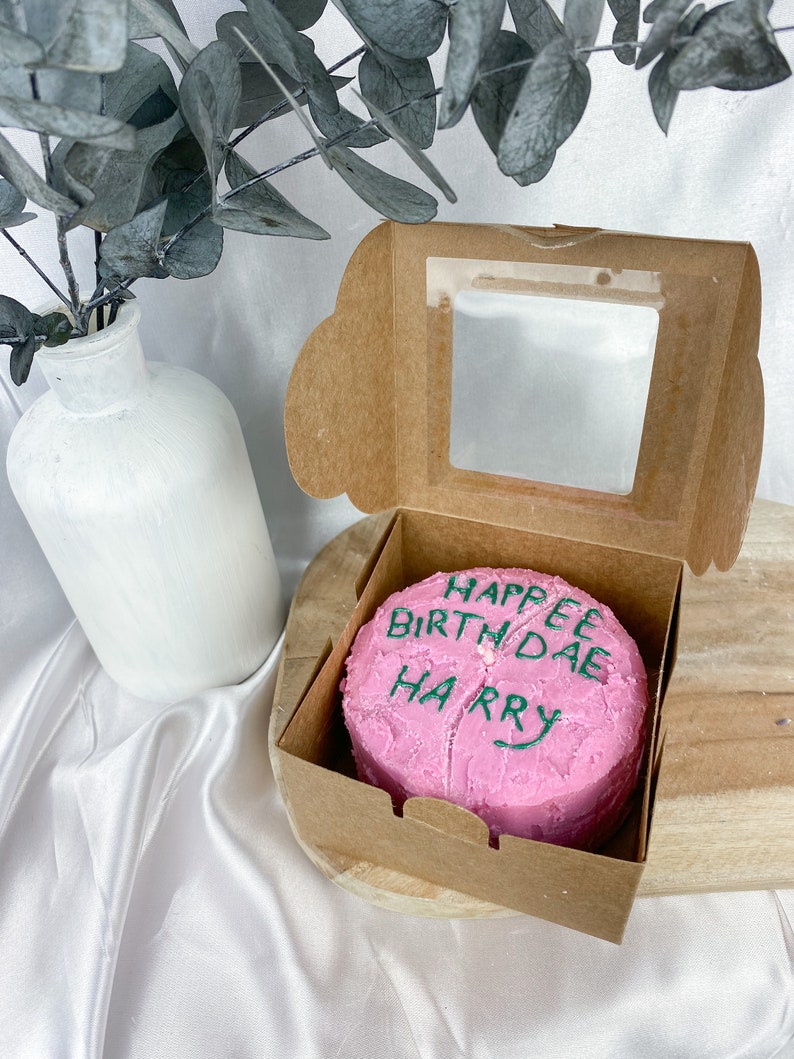 May include: A pink cake with green icing that says "Happy Birthday Harry" in a cardboard box with a clear window.