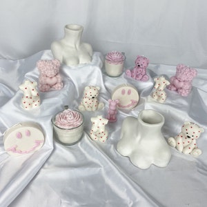 May include: Assortment of pink and white scented candles in various shapes, including bears, flowers, and smiley faces. The candles are arranged on a white surface.