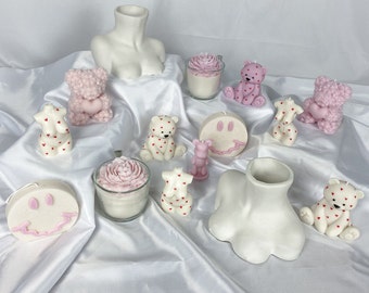 May include: Assortment of pink and white scented candles in various shapes, including bears, flowers, and smiley faces. The candles are arranged on a white surface.