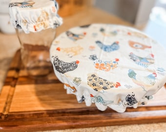 May include: Two reusable food covers with a white background and a colorful chicken pattern. The covers are made of fabric and have a drawstring closure.