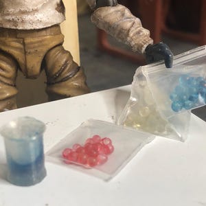 Puede incluir: Una escena en miniatura con un vaso de pl&aacute;stico transparente con l&iacute;quido azul, tres peque&ntilde;as bolsas de pl&aacute;stico que contienen objetos esf&eacute;ricos amarillos, rojos y azules, y una figura con ropa beige. Los objetos est&aacute;n sobre una superficie blanca.