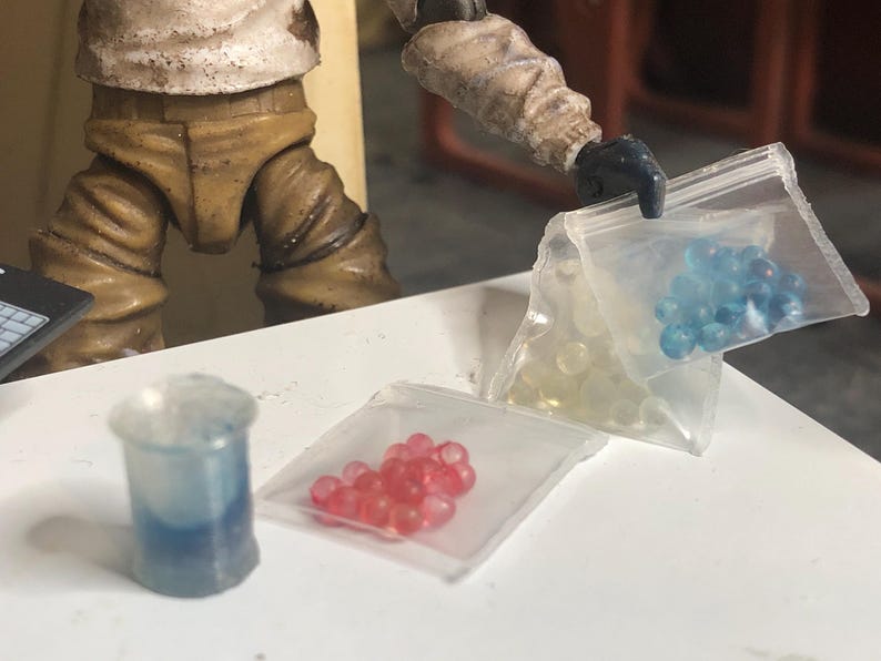 Puede incluir: Una escena en miniatura con un vaso de pl&aacute;stico transparente con l&iacute;quido azul, tres peque&ntilde;as bolsas de pl&aacute;stico que contienen objetos esf&eacute;ricos amarillos, rojos y azules, y una figura con ropa beige. Los objetos est&aacute;n sobre una superficie blanca.
