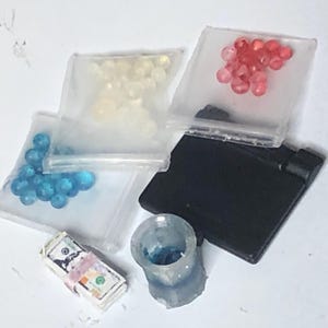 Puede incluir: Un surtido de art&iacute;culos en miniatura, que incluyen peque&ntilde;as bolsas llenas de cuentas azules, blancas y rojas. Tambi&eacute;n hay una pila de dinero de juguete, un rect&aacute;ngulo negro y un peque&ntilde;o recipiente transparente con contenido azul.