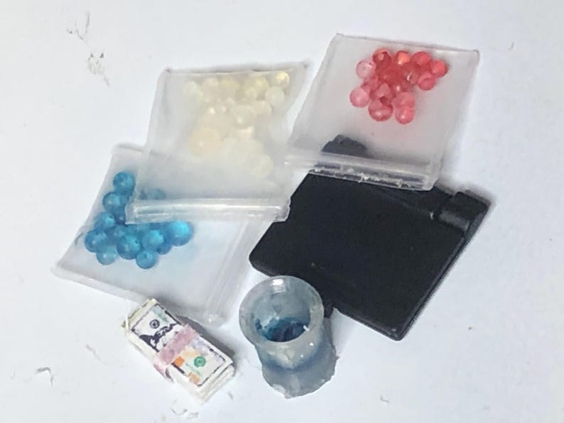 Puede incluir: Un surtido de art&iacute;culos en miniatura, que incluyen peque&ntilde;as bolsas llenas de cuentas azules, blancas y rojas. Tambi&eacute;n hay una pila de dinero de juguete, un rect&aacute;ngulo negro y un peque&ntilde;o recipiente transparente con contenido azul.