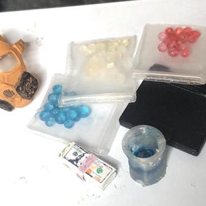 Puede incluir: Un surtido de objetos en miniatura, que incluyen una m&aacute;scara de gas marr&oacute;n y beige, peque&ntilde;as bolsas de cuentas de varios colores, un objeto rectangular negro, una peque&ntilde;a taza transparente y un fajo de dinero de juguete.