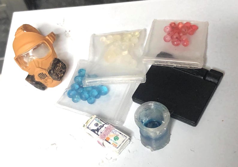 Puede incluir: Un surtido de objetos en miniatura, que incluyen una m&aacute;scara de gas marr&oacute;n y beige, peque&ntilde;as bolsas de cuentas de varios colores, un objeto rectangular negro, una peque&ntilde;a taza transparente y un fajo de dinero de juguete.