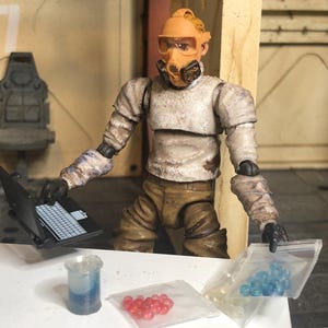 Puede incluir: Figura de una persona con traje de protecci&oacute;n con un port&aacute;til, un vaso de precipitados y bolsas de cuentas rojas y azules. La figura lleva una m&aacute;scara amarillo-naranja y est&aacute; en una habitaci&oacute;n con una silla y el n&uacute;mero 7.