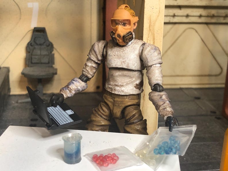 Puede incluir: Figura de una persona con traje de protecci&oacute;n con un port&aacute;til, un vaso de precipitados y bolsas de cuentas rojas y azules. La figura lleva una m&aacute;scara amarillo-naranja y est&aacute; en una habitaci&oacute;n con una silla y el n&uacute;mero 7.