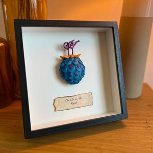Peut inclure: Une œuvre d'art encadrée représentant une sculpture 3D bleue et violette d'un fruit avec une tige et des feuilles dorées. Le texte "Uo Uo no Mi Kaido" est écrit sur un morceau de parchemin en dessous de la sculpture.