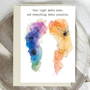 May include: Greeting card with watercolor wings in shades of blue, purple, orange, and yellow. The wings have black constellation lines and dots. The text reads, "Your light meets mine, and everything feels possible."