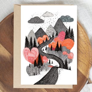 May include: A greeting card featuring a whimsical landscape illustration. The artwork depicts a winding road through a forest with heart-shaped trees in shades of red and orange, set against a backdrop of mountains and clouds. The card is paired with a brown envelope.