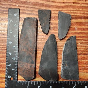 May include: A collection of dark, irregularly shaped stone pieces, possibly flakes or shards, arranged on a wooden surface. A ruler is placed next to the stones for scale, showing measurements in inches. The stones vary in size and color, with some displaying a reddish-brown hue.