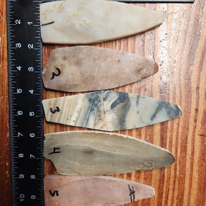 May include: Five elongated, pointed stone artifacts arranged vertically next to a black L-shaped ruler. The stones are numbered 1 through 5 in black ink. They feature varied textures and colors, including cream, tan, grey, and brown, with lengths ranging from approximately 2.5 to 3.5 inches.
