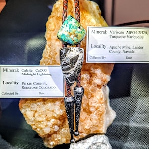 May include: A bolo tie featuring a turquoise and black and white stone pendant, with a brown leather cord and decorative silver tips. The image includes labels with mineral information: Calcite and Variscite.