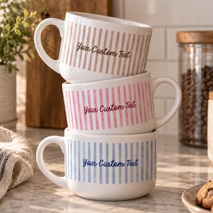 May include: A stack of three white ceramic mugs with handles, each featuring vertical stripes in light brown, pink, or blue. Each mug has the text Your Custom Text printed in a matching color script font on the front center of the exterior surface.