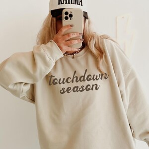 Peut inclure: Un sweat-shirt à col rond couleur sable avec le texte marron foncé touchdown season imprimé sur la poitrine. Le vêtement est associé à un jean bleu clair délavé et une casquette de baseball crème avec le mot KARMA en texte noir.