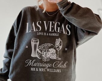 May include: A dark grey sweatshirt with a white graphic print that says "Las Vegas Love is a Gamble" and features a beer, playing cards, dice, and a cocktail. The graphic also says "Marriage Club Mr. & Mrs. Williams".