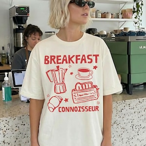 May include: Cream-colored t-shirt with the words "BREAKFAST CONNOISSEUR" in red, along with red illustrations of a coffee pot, cup, toaster, and croissant. The shirt is worn by a person wearing sunglasses.