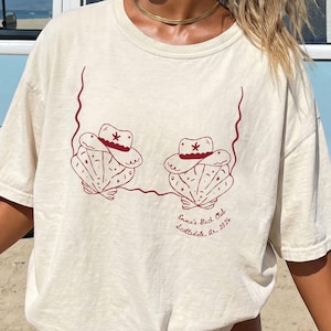 Puede incluir: Camiseta de color marfil con un diseño gráfico rojo oscuro. El diseño presenta dos formas de conchas marinas con sombreros de vaquero y el texto "Emma's Bach Club, Scottsdale, Ar. 2026".