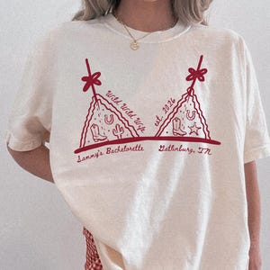 May include: Ivory-colored t-shirt with a dark red graphic design. The design features a bra-like shape with text that reads "Wild Wild Wife est. 2026" and "Sammy's Bachelorette Gatlinburg, TN". The shirt is paired with red and white checkered shorts.