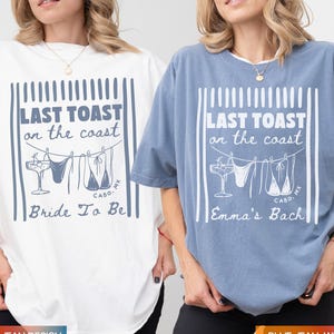 Op de afbeelding: Twee oversized t-shirts, één wit met blauwe opdruk en één blauw met witte opdruk. De afbeelding toont verticale strepen, een cocktailglas en badkleding aan een waslijn met de tekst LAST TOAST on the coast, CABO, MX, Bride To Be en Emma's Bach.