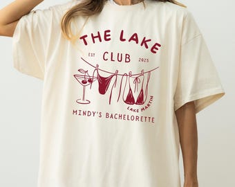 Op de afbeelding: Ivoor kleurig T-shirt met een donkerrode afbeelding. Het ontwerp bevat de tekst "THE LAKE CLUB EST 2025" boven een lijn met een martiniglas en hangende badpakken. Daaronder staat "MINDY'S BACHELORETTE LAKE MARTIN."