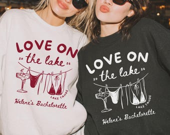 Op de afbeelding: Twee vrouwen dragen sweatshirts met de tekst "LOVE ON the lake 26" en "Helene's Bachelorette" erop. De sweatshirts hebben een afbeelding van een cocktailglas en badpakken. Eén sweatshirt is wit, de andere is zwart.