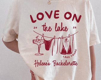 Op de afbeelding: Crèmekleurig T-shirt met een rode afbeelding met de tekst "LOVE ON the lake" met illustraties van een cocktail en lingerie. De tekst "Helene's Bachelorette" staat ook op het shirt. Het shirt wordt gecombineerd met een rood-witte geruite short.