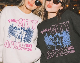 May include: Two people wearing sunglasses and sweatshirts. The sweatshirts are white and black, with the text "PARK CITY APRES SKI CLUB" in pink and blue. The design includes a skier, trees, and mountains.