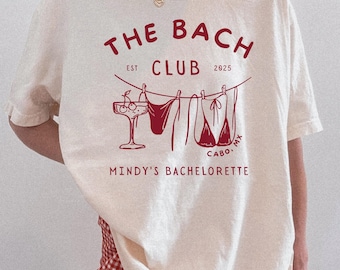 May include: An ivory t-shirt featuring a dark red graphic print that reads THE BACH CLUB EST 2025, CABO, MX, and MINDY'S BACHELORETTE. The design includes a line drawing of a cocktail glass and two bikini pieces hanging from a clothesline.