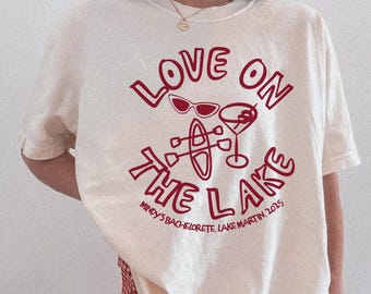 May include: Ivory t-shirt with a red graphic design that says "Love on the Lake" with a martini glass, sunglasses, and a canoe. The text below the graphic says "Mindy's Bachelorette, Lake Martin, 2025".